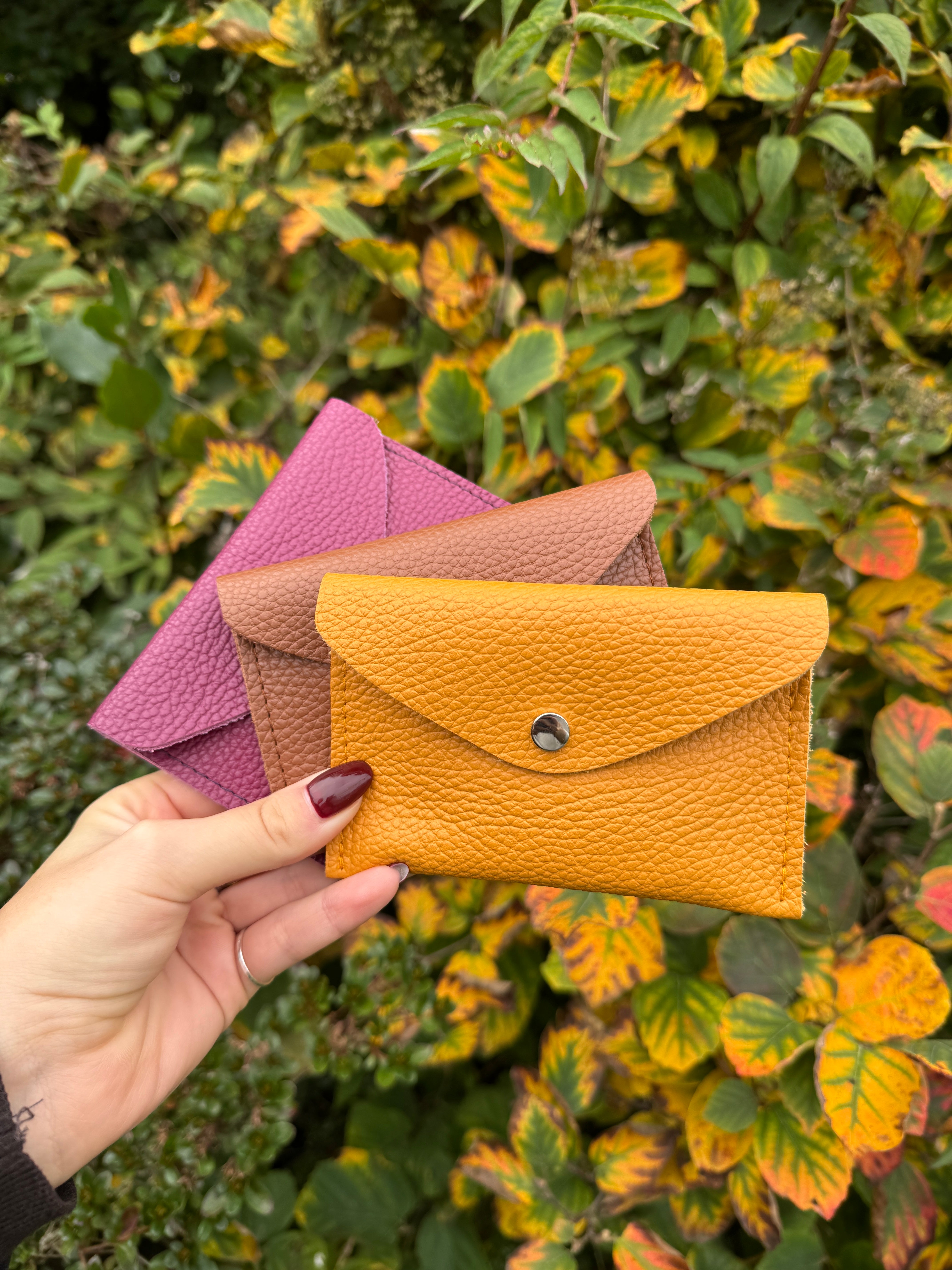 Hand holding three small leather pouches in front of a leafy green background