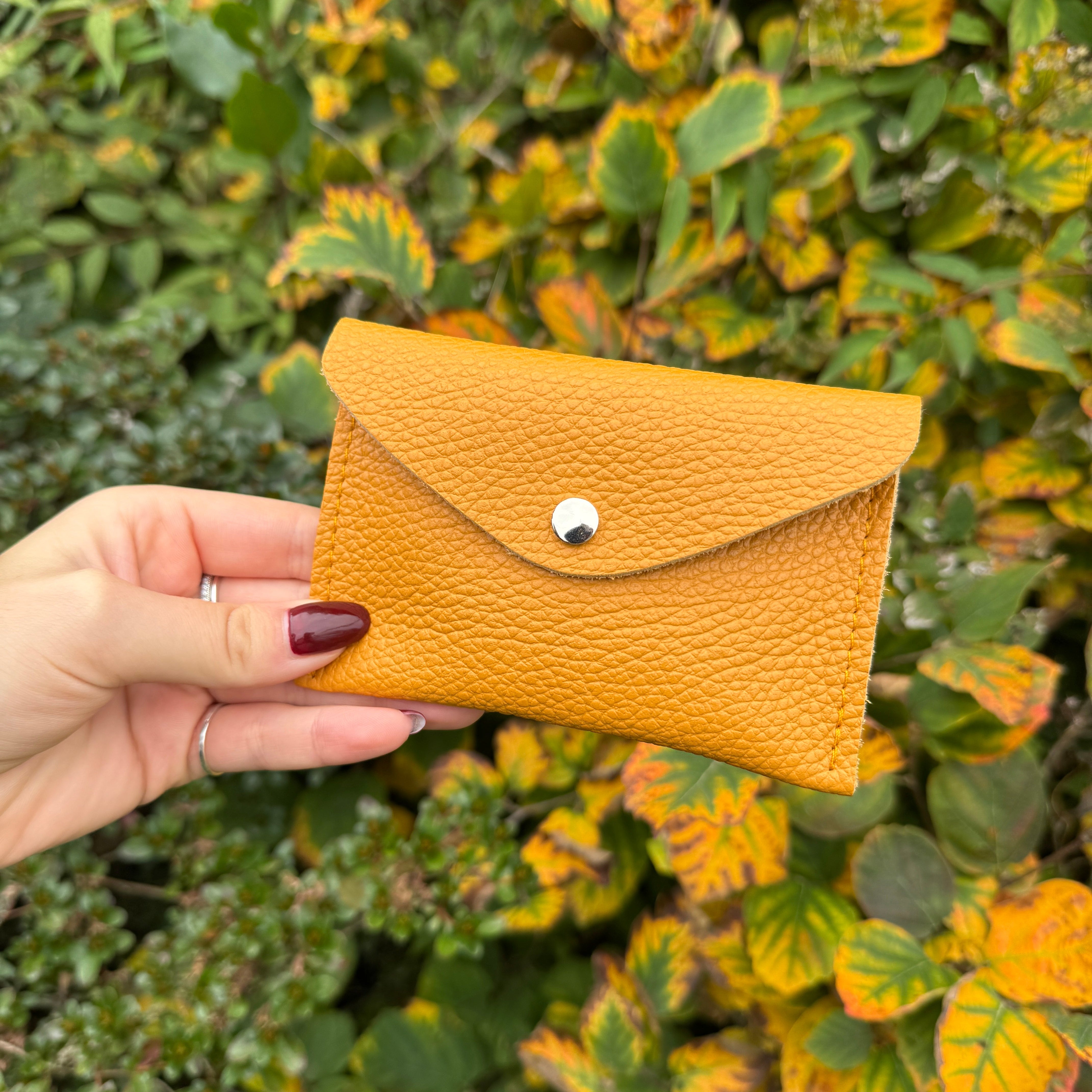 Hand holding a pumpkins spice orange leather cardholder against a green leafy background