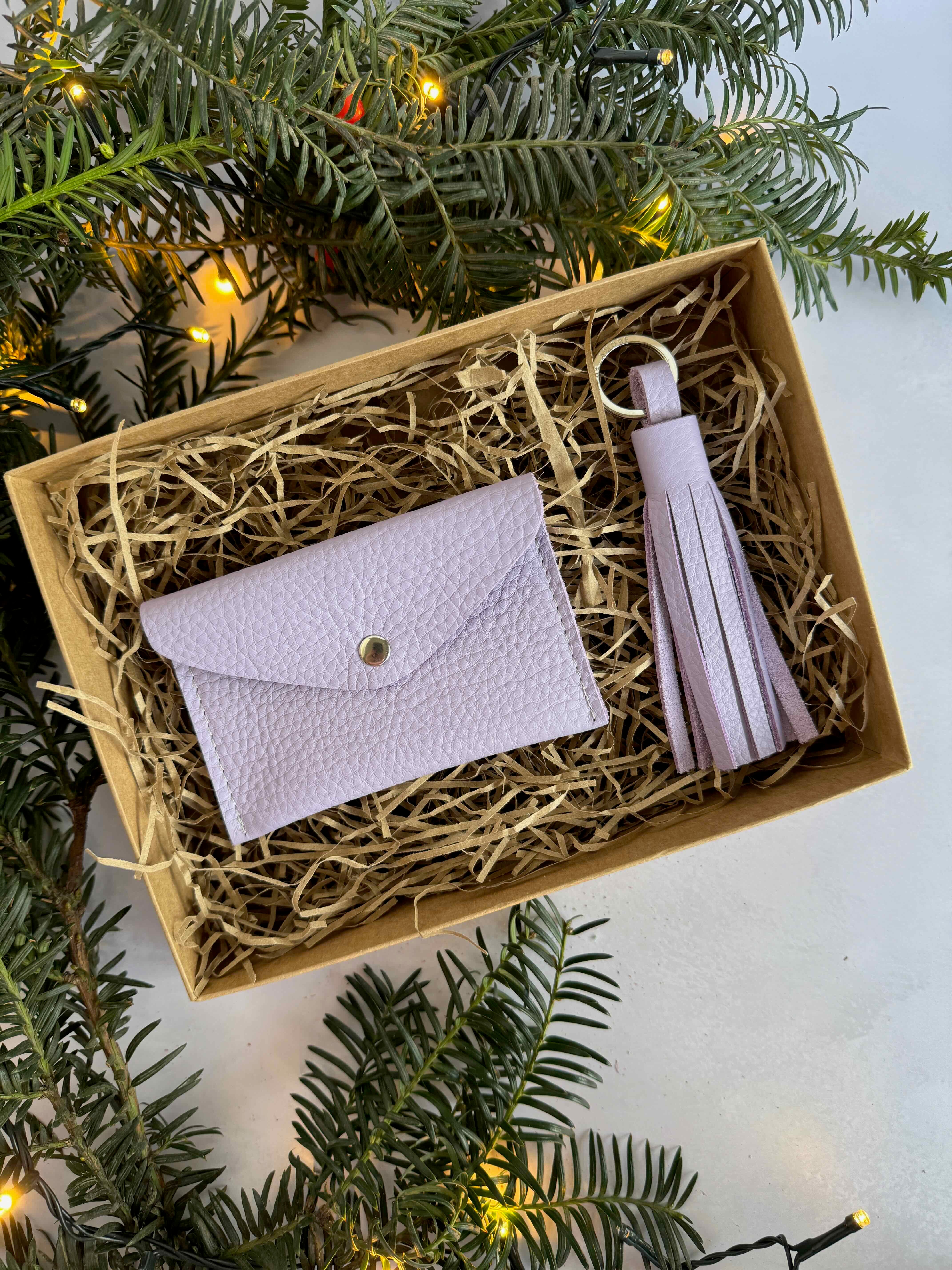 Gift set with a lilac wallet and tassel in a decorative box, surrounded by Christmas tree branches.