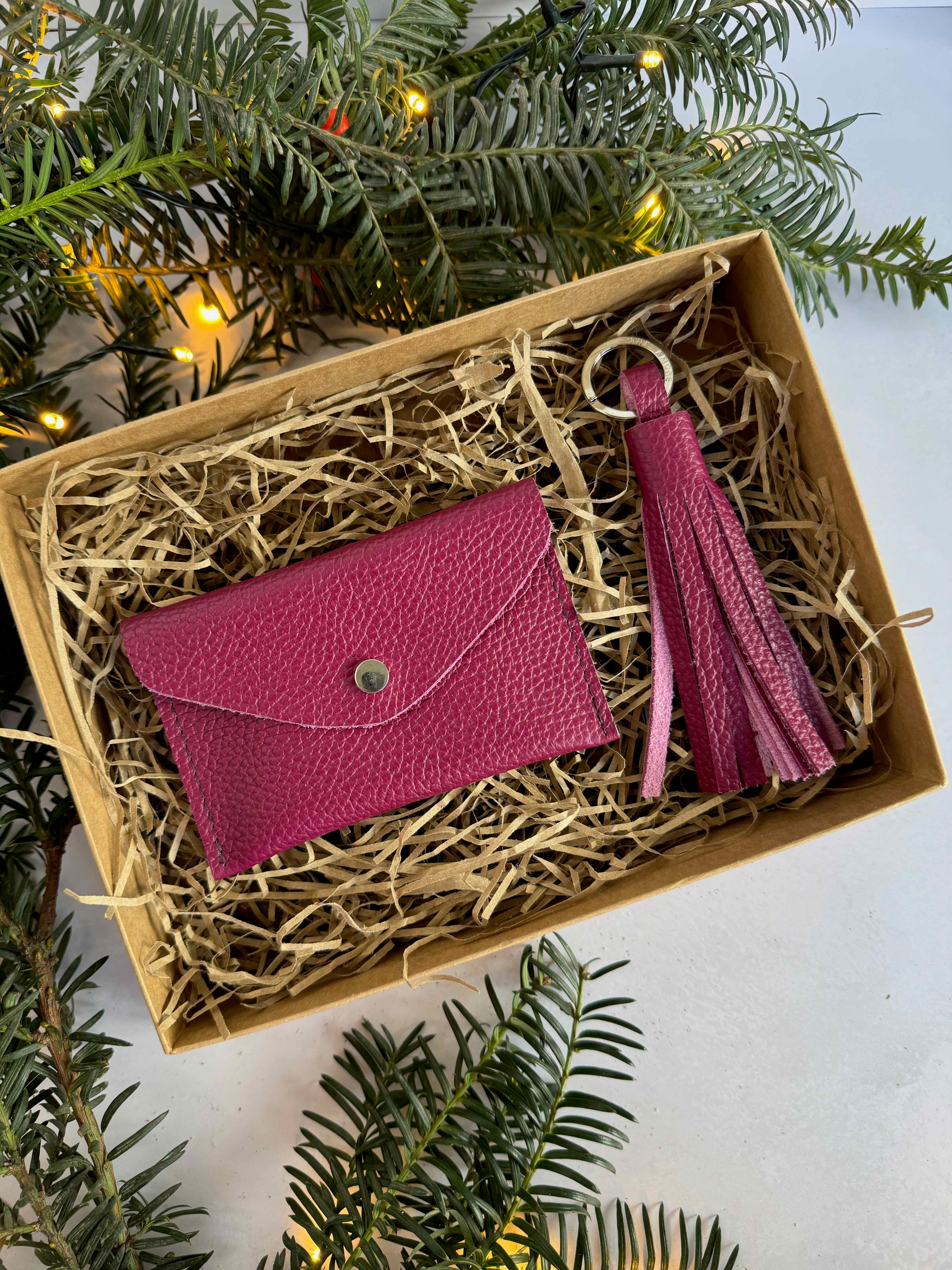 plum leather wallet and keychain with tassel in a decorative box surrounded by greenery.