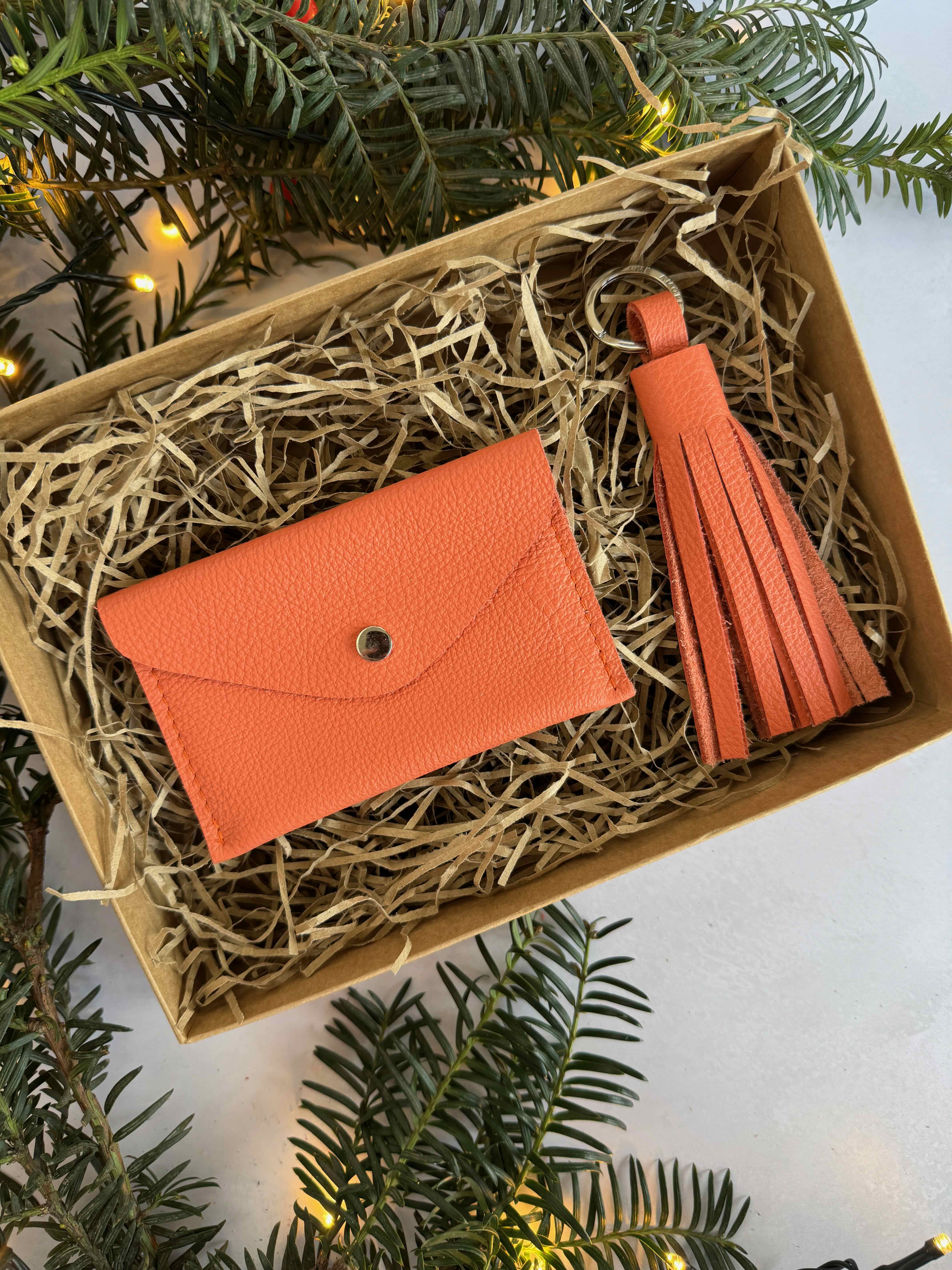 Orange wallet and tassel keychain in a box with Christmas greenery around.
